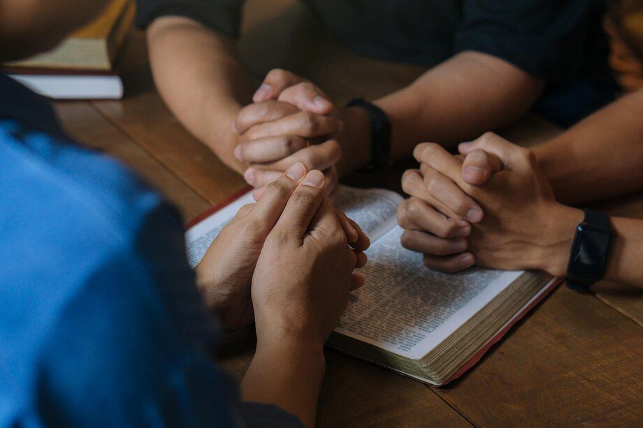 Three people praying with hands clasped over an open Bible on a wooden table.