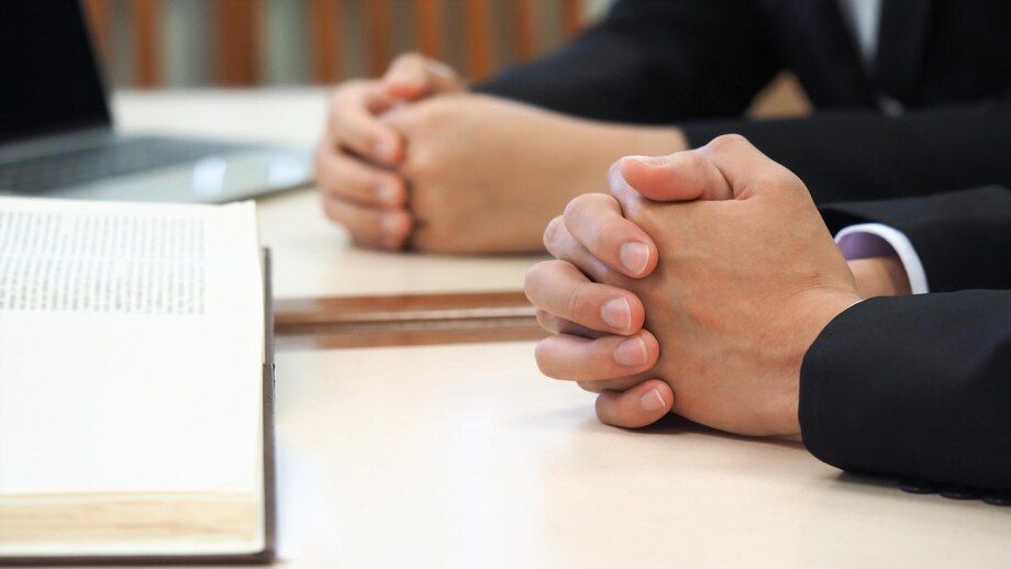 Hands clasped on a table, likely in a meeting or consultation, near a book and a laptop.