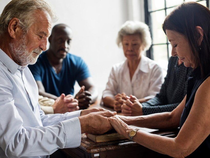 Group of people with hands clasped in prayer around a table.