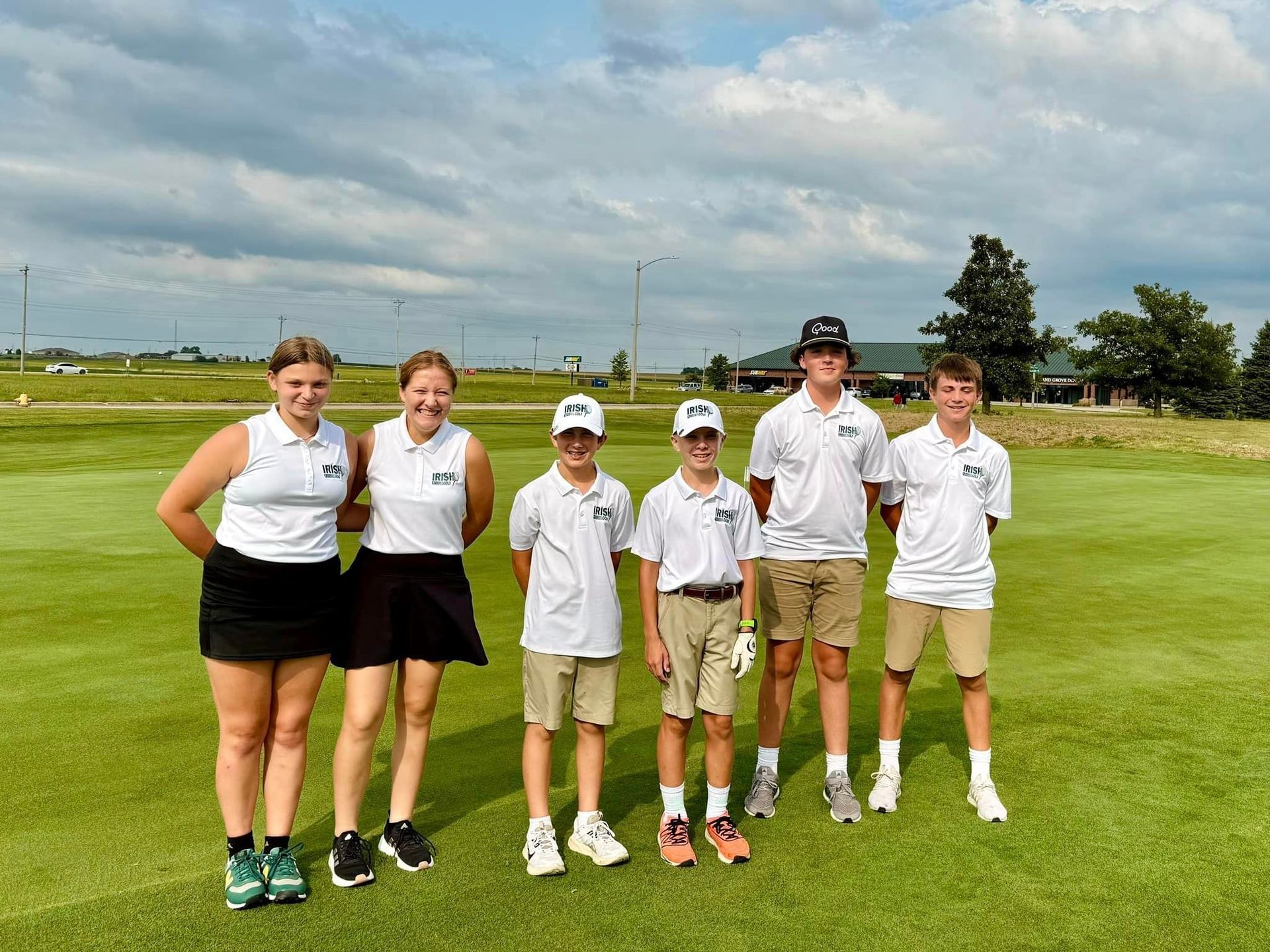 Golf team poses on a green. White shirts, khaki shorts, cloudy sky, golf course background.