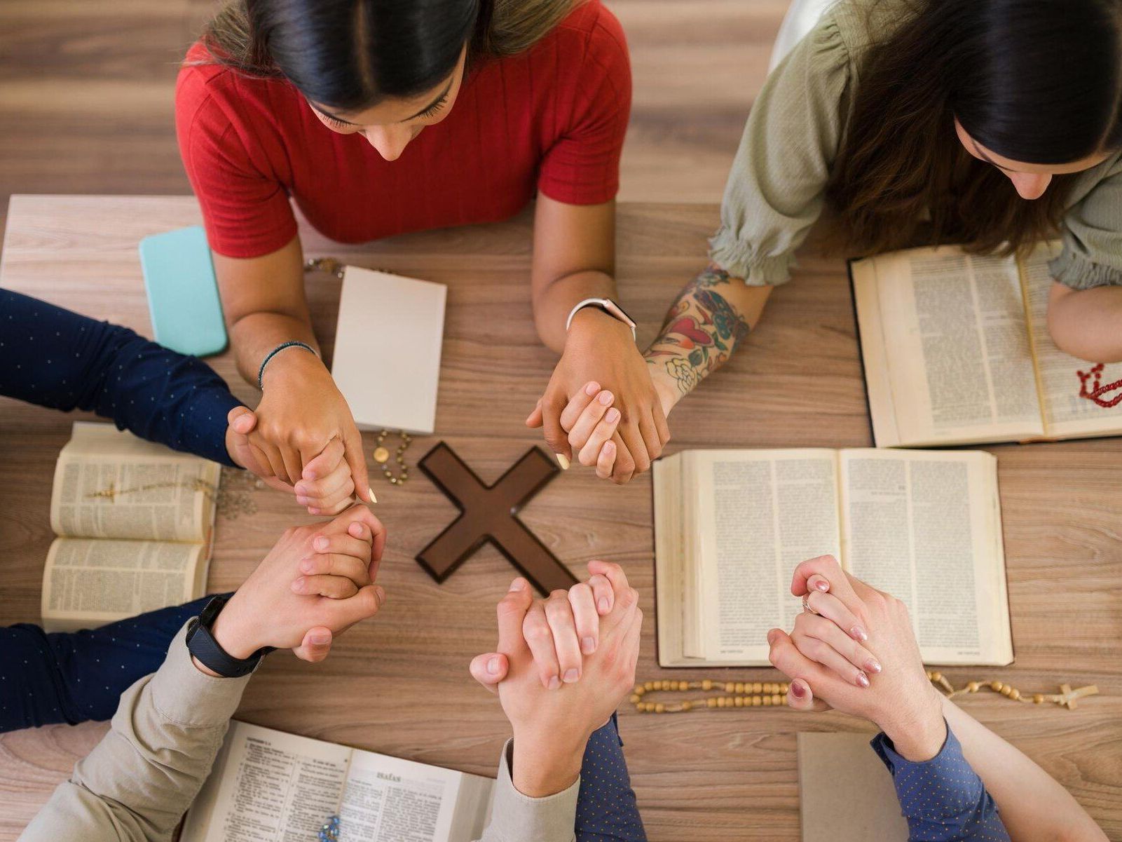 People praying around a wooden cross and open Bibles on a table, holding hands.