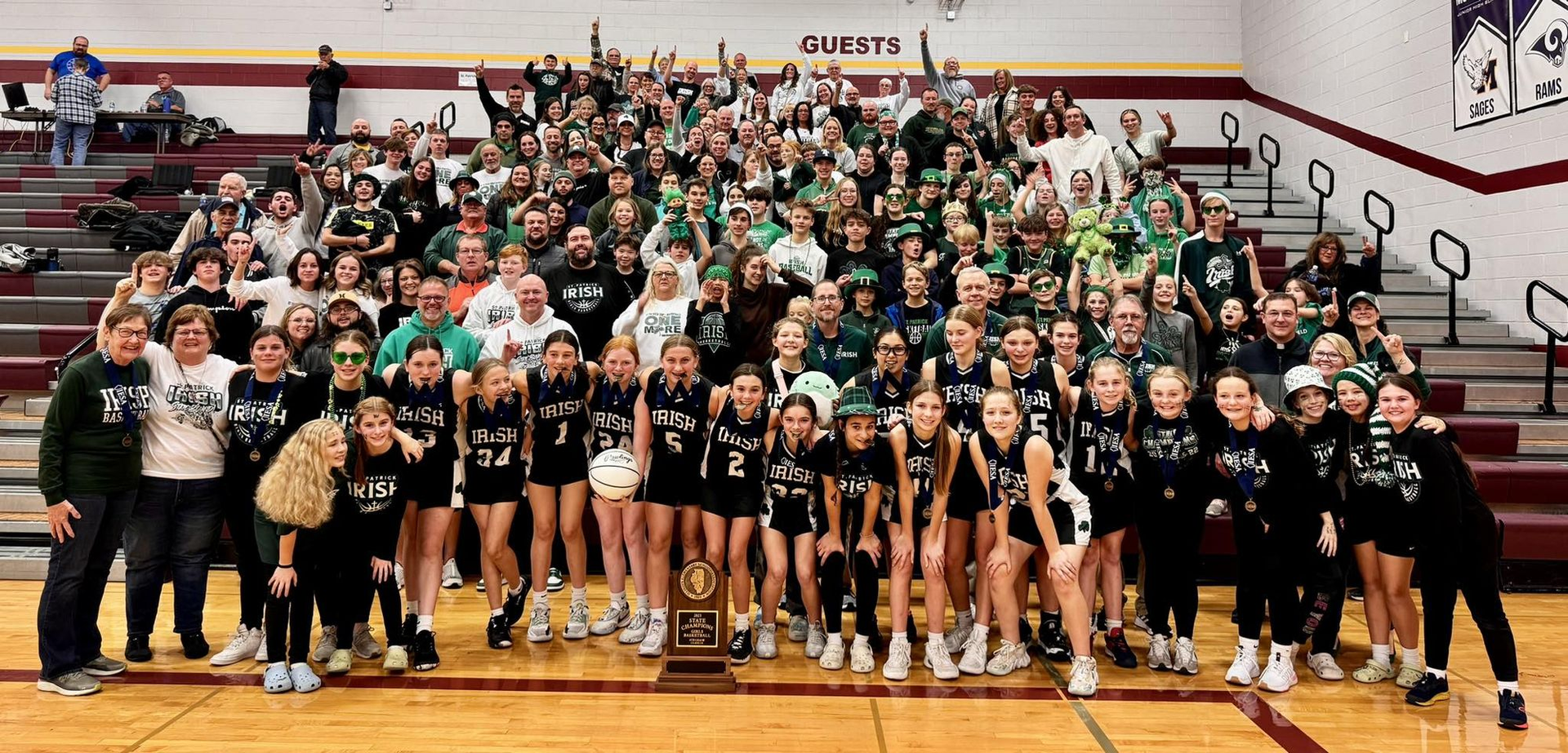 A large group of people, many wearing green and black, pose in a gymnasium, holding a trophy and celebrating.