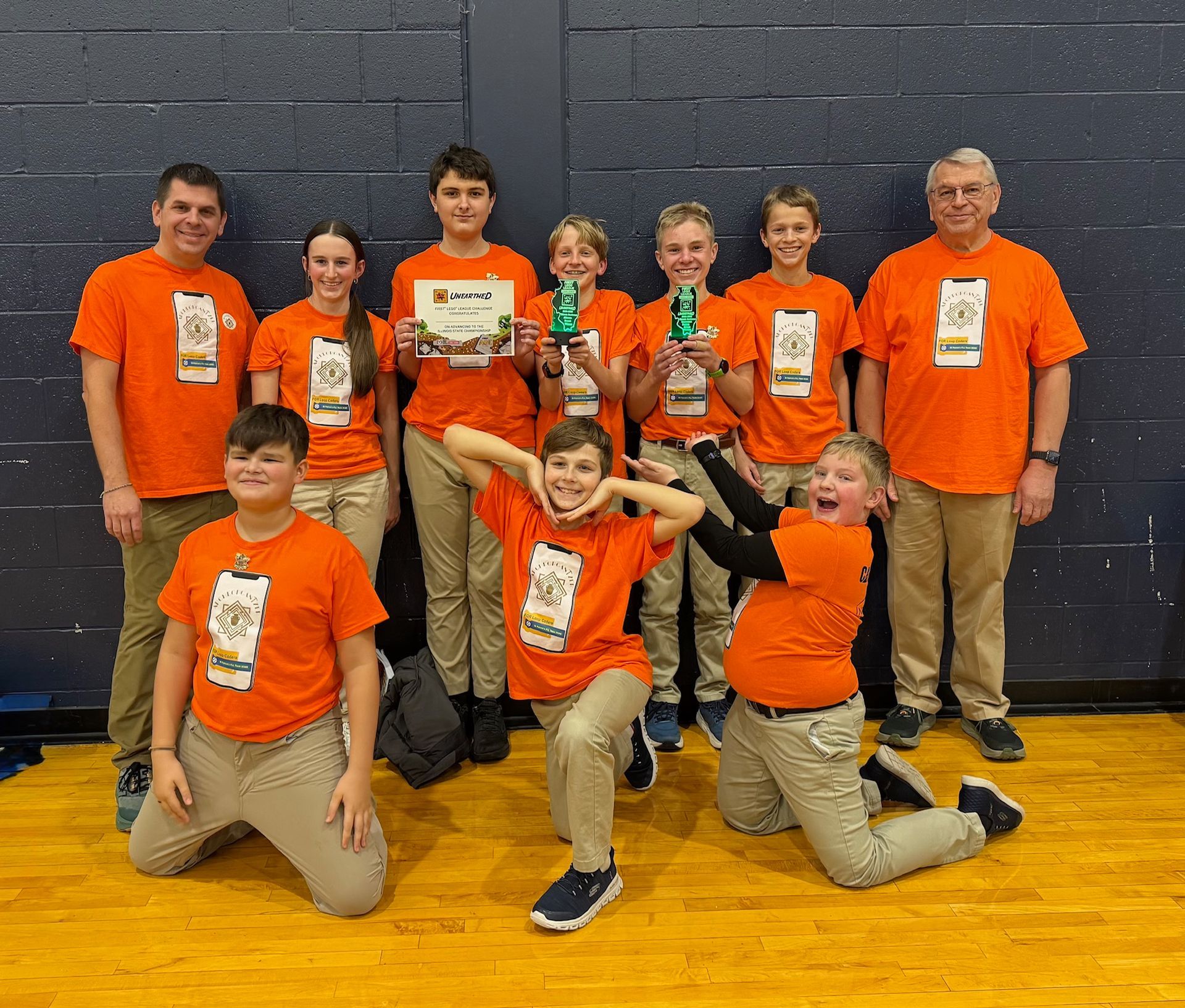 Group of people in orange shirts posing in a gym, some kneeling and holding trophies.