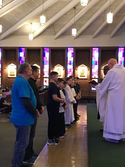 People in a church service: men, boys in white robes, a priest with book, near stained glass.