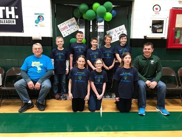 Group of children and adults wearing matching shirts posing by green balloons.