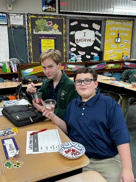 Two boys in a classroom hold snacks. One smiles, the other looks at the camera. Classroom setting with posters.