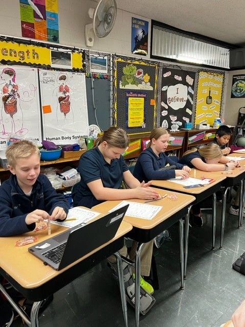 Students in classroom working at desks, using papers and a laptop.