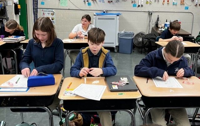 Students at desks in a classroom are focused on a school project. Some are wearing blue uniform shirts.