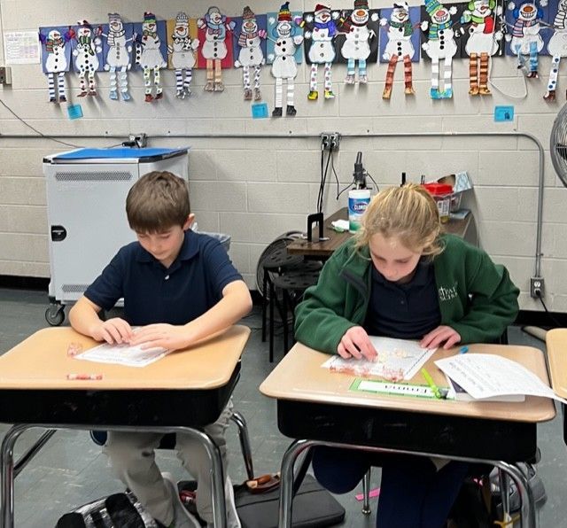 Two students working at desks in a classroom; decorated with snowman cutouts.