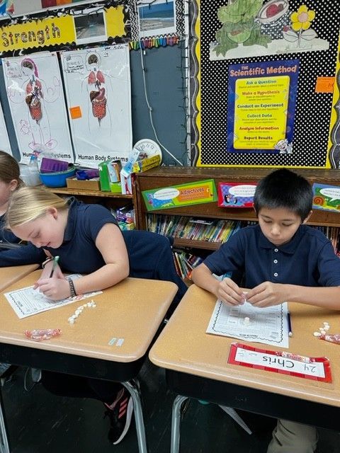 Two students in blue shirts at desks; one writes, the other works with small objects. Classroom setting.