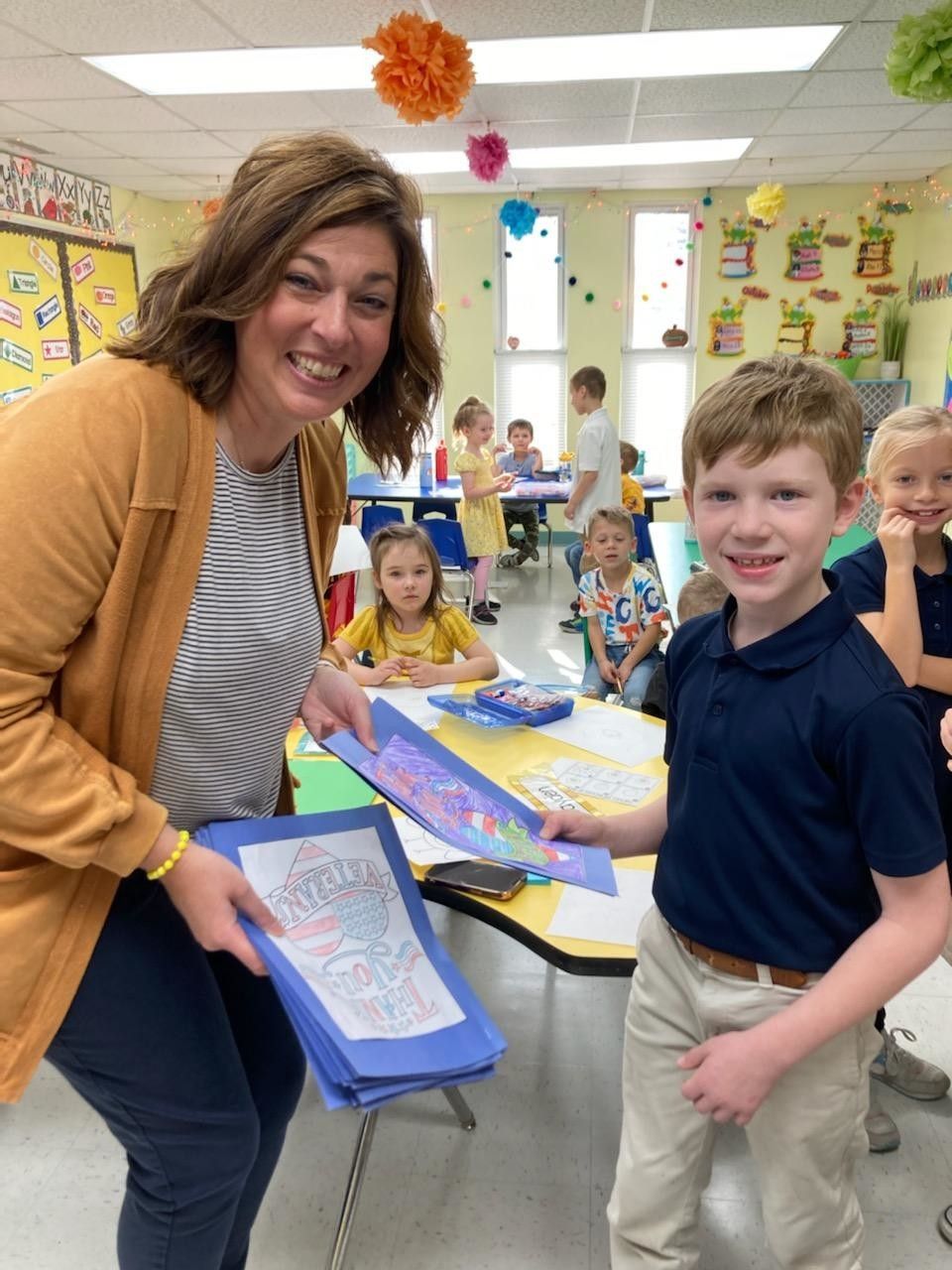 Teacher smiling with student holding artwork in classroom. Other students are in the background.