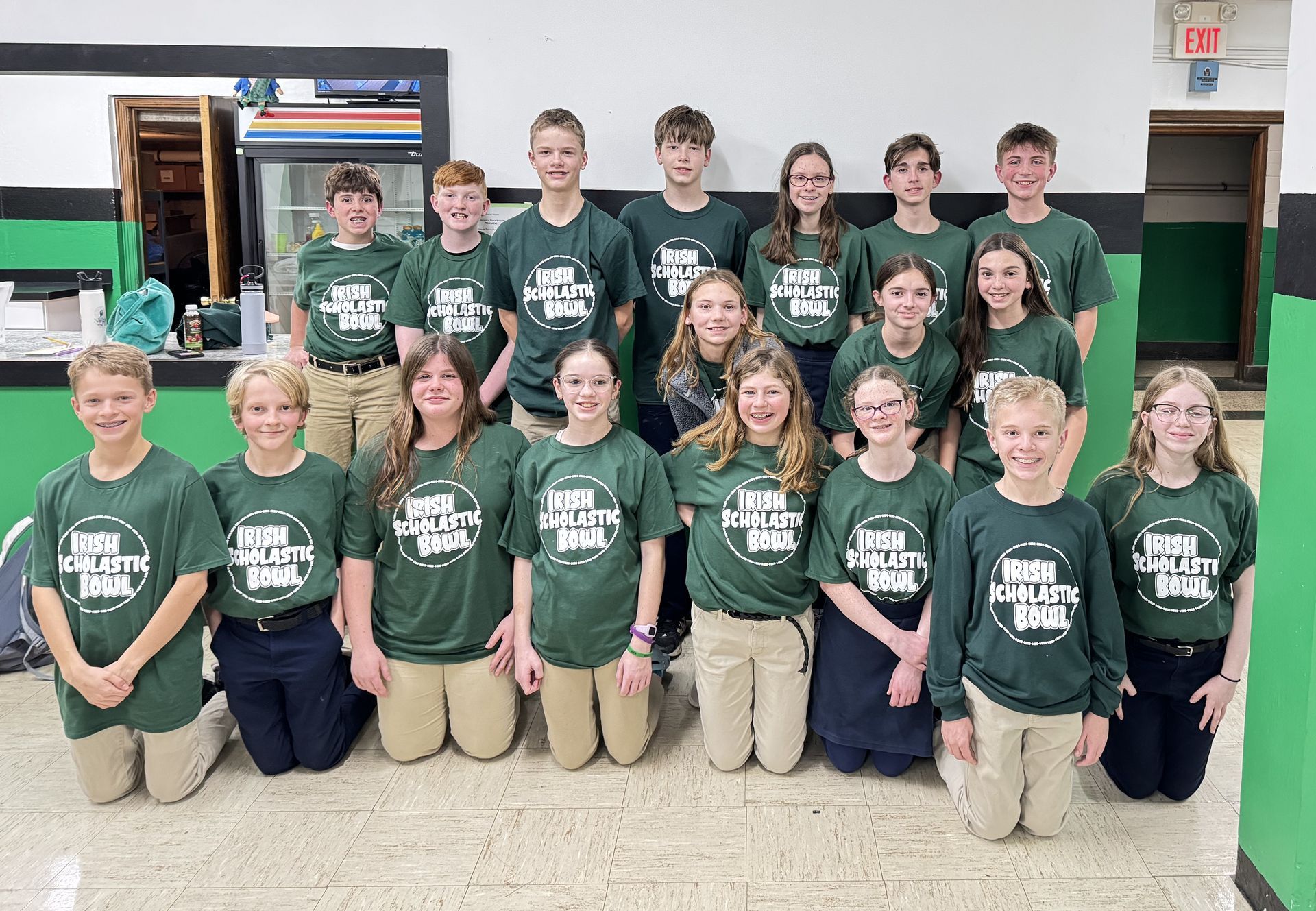 Group of teens in green shirts with a logo, posing indoors. Some kneel, all smile.