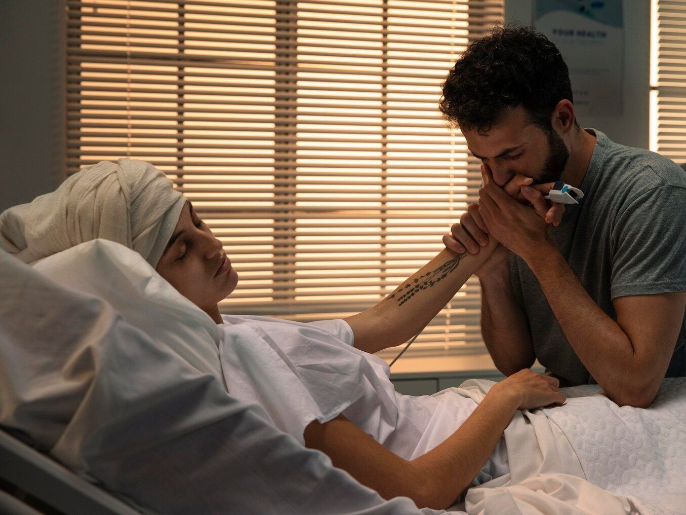 Man comforts woman in hospital bed, kissing her hand. Woman has head wrapped, possibly ill. Sunlit room.