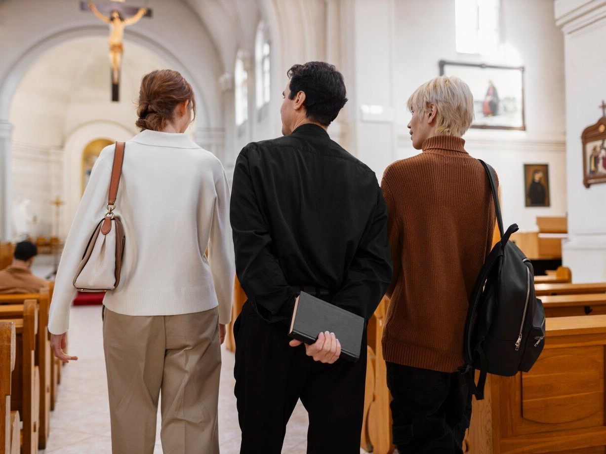 Three people walking in a church, one carrying a bible. Backs to the viewer, toward a crucifix and altar.
