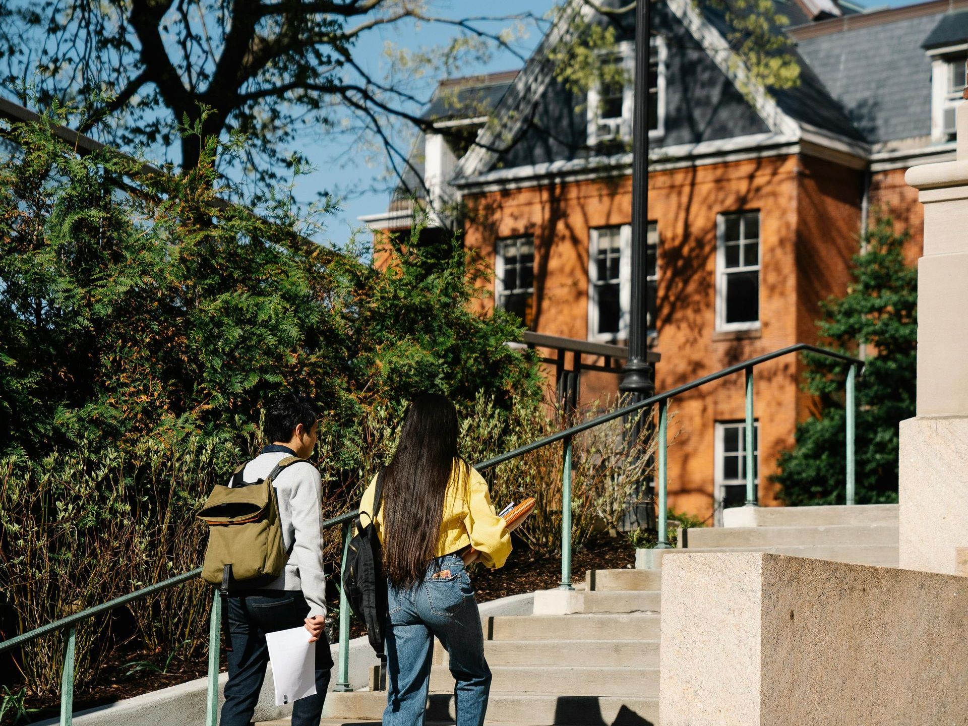Two students walk up stairs toward a brick building. One carries a backpack, the other a yellow shirt. Outdoors.