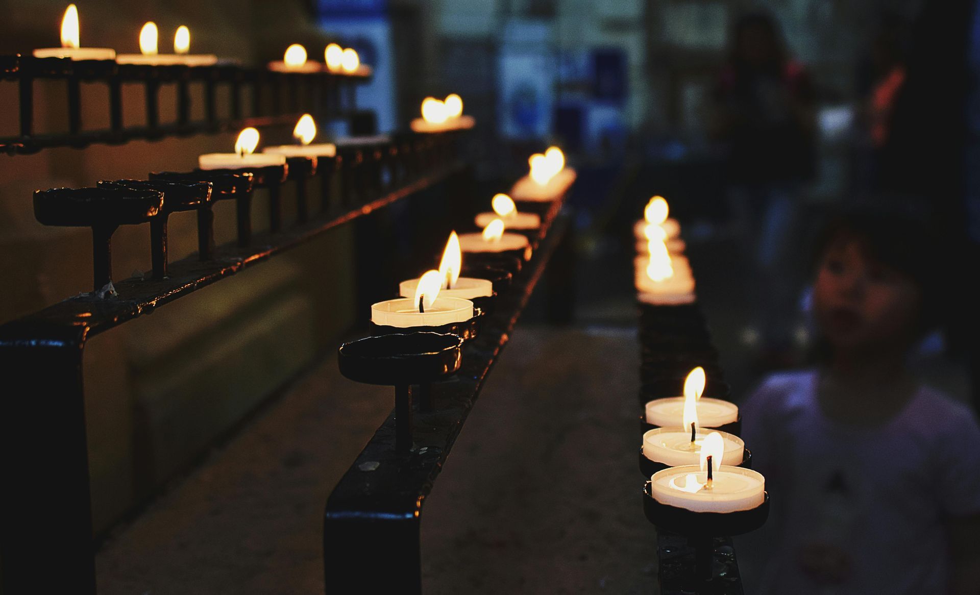 Rows of lit candles on a black rack, inside a dimly lit building. A child looks on.