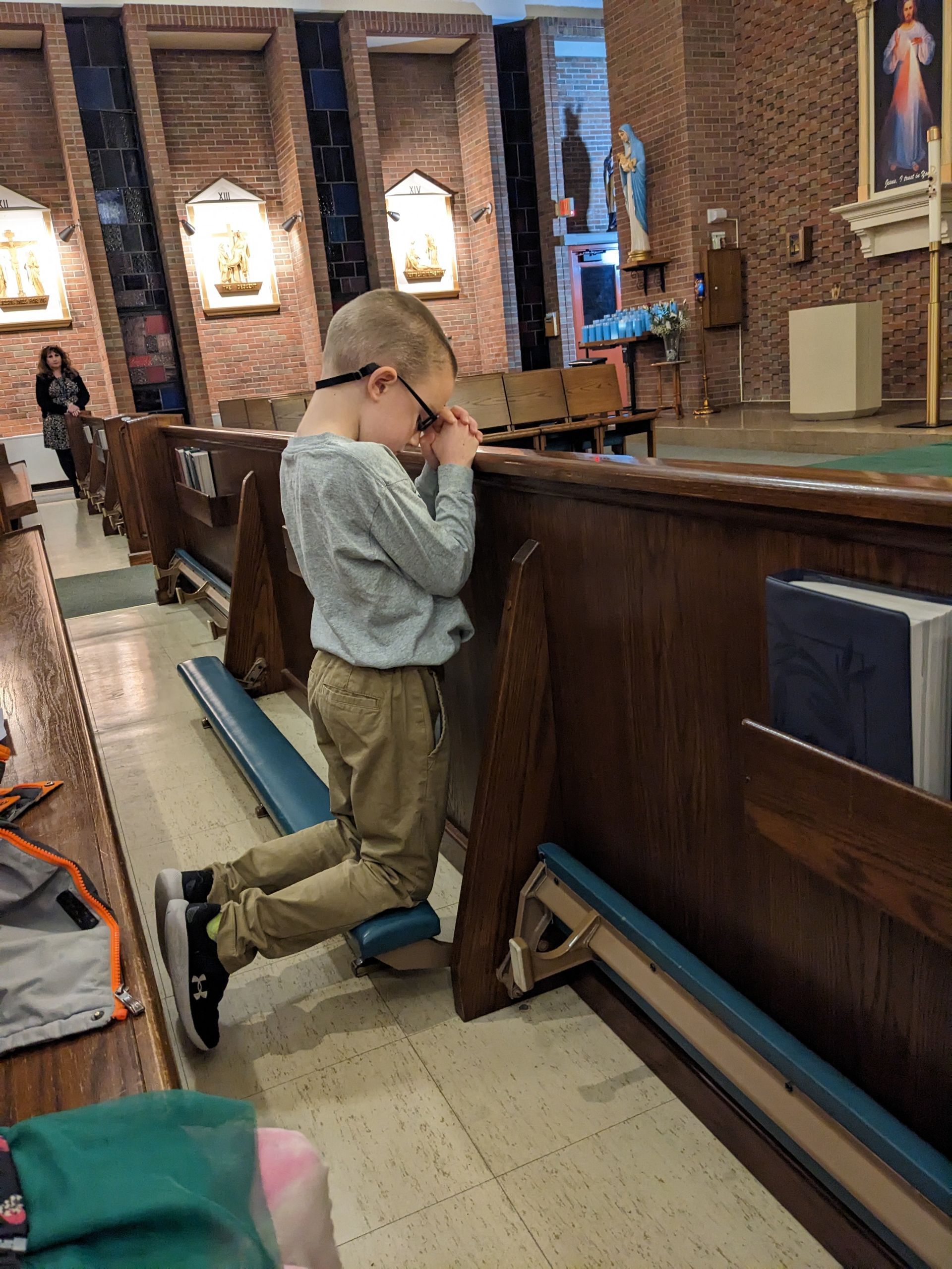 Young boy kneeling and praying in a church, hands clasped, head bowed. Wooden pews and stained-glass windows.