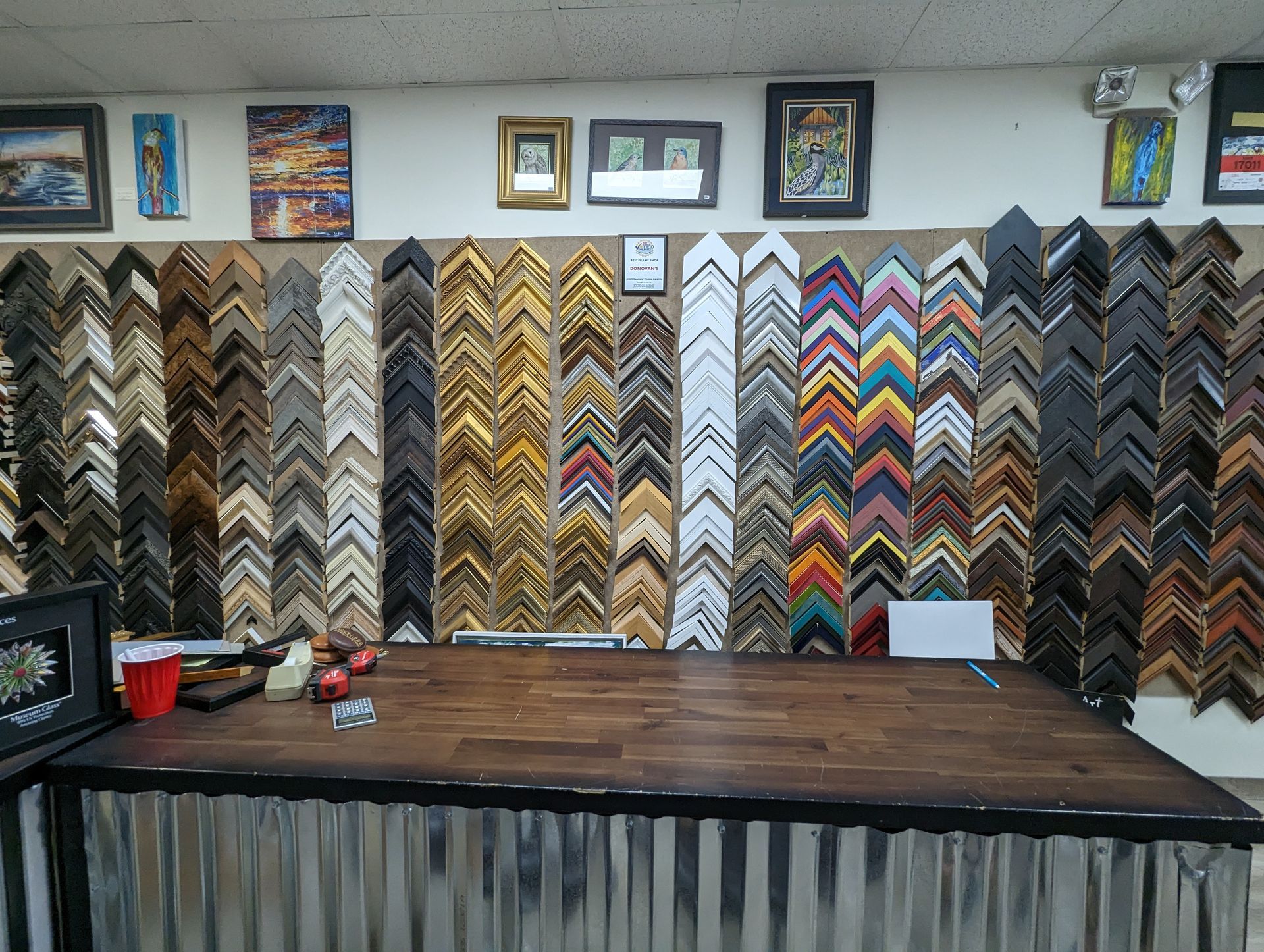 Display of picture frames in a framing shop, showcasing various styles and colors against a white wall.