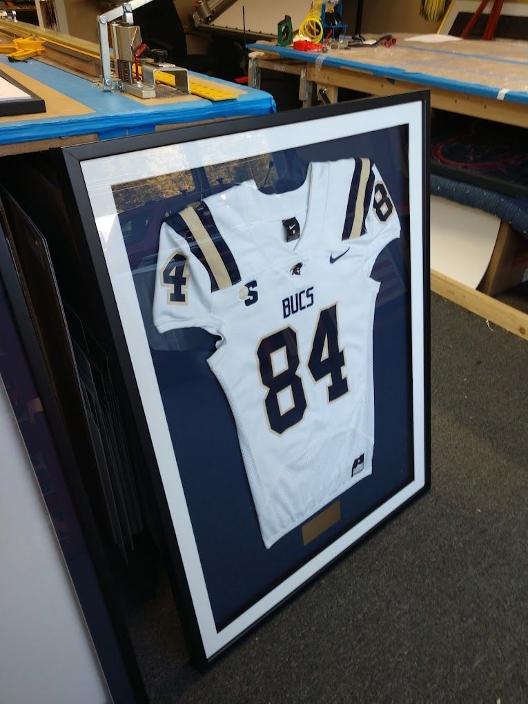White football jersey with navy and gold details in a black frame, on a blue background.
