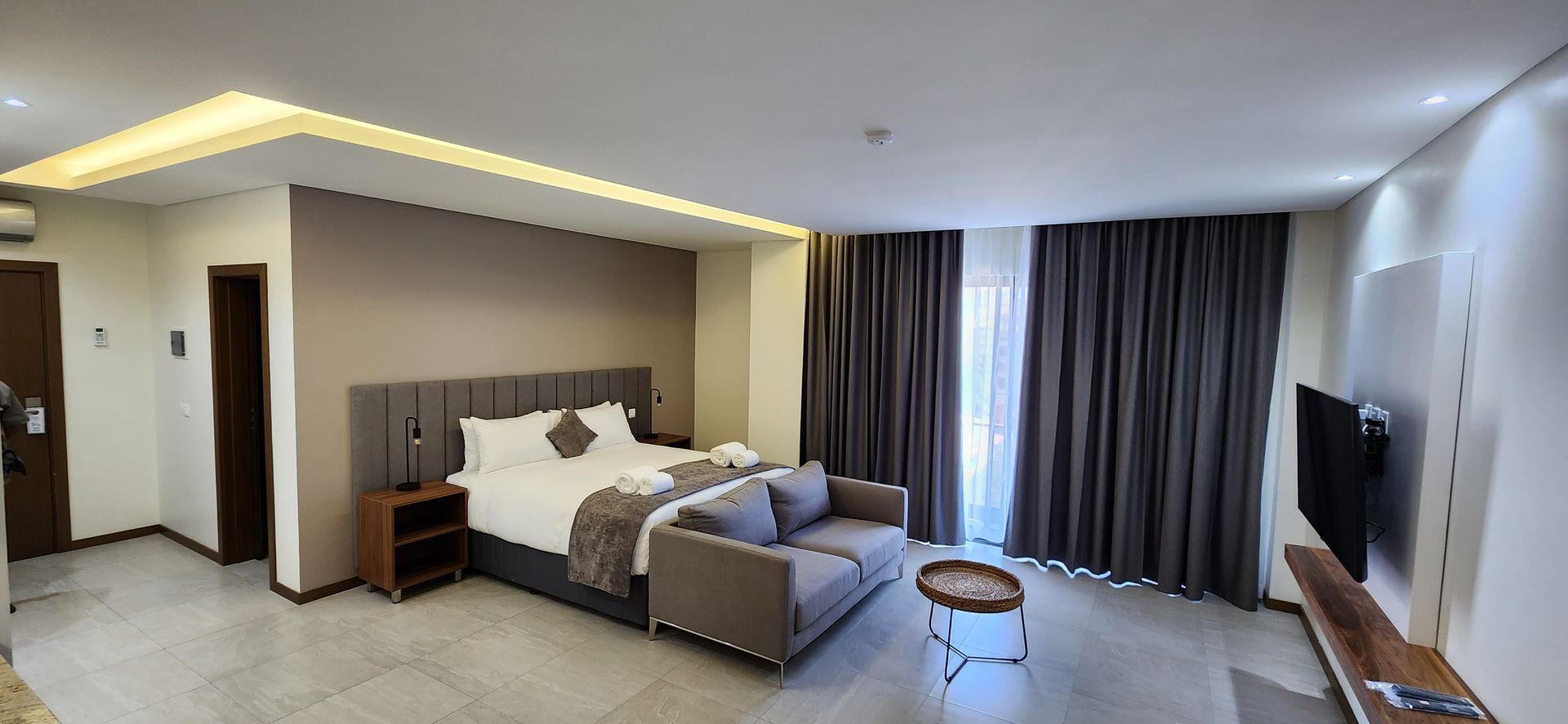 Hotel room with bed, sofa, curtains, and wall-mounted TV. Neutral color palette and modern design.