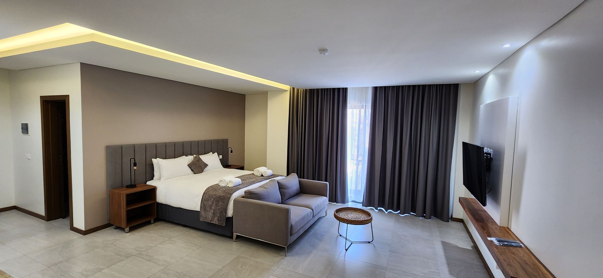 A modern hotel room with a bed, sofa, TV, and large window with gray curtains.