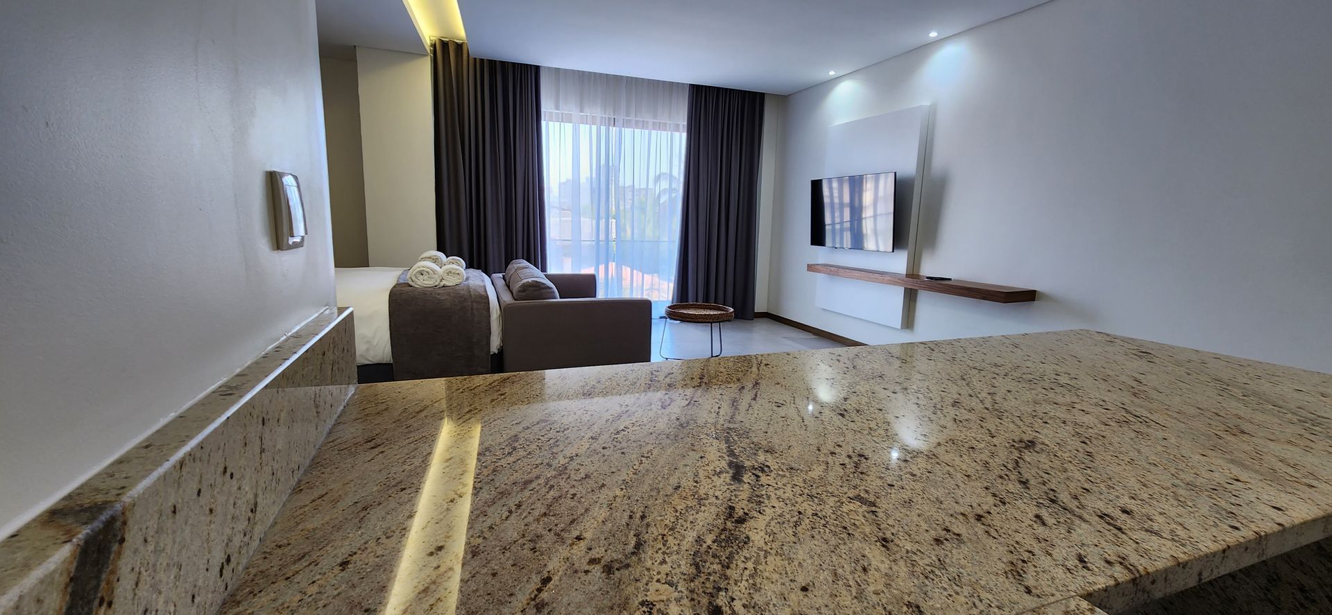 Modern living room with a granite countertop, sofa, TV, and large window with curtains.