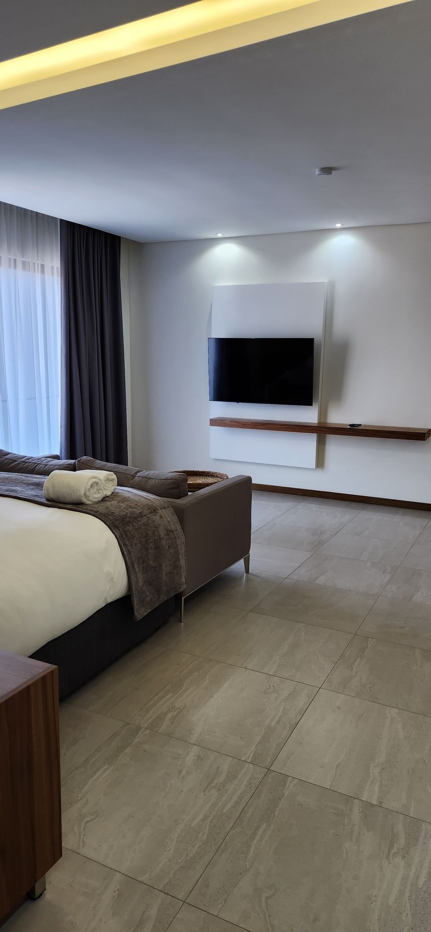 Modern bedroom with a bed, TV on the wall, gray floor tiles, and window with curtains.