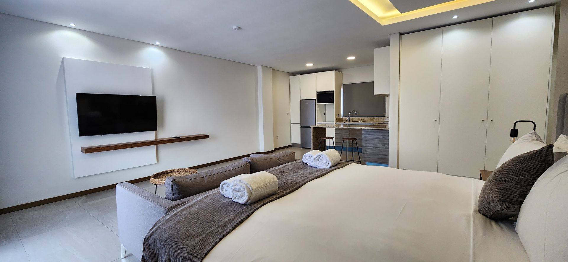 Modern studio apartment with a bed, sofa, TV, and kitchen area; white walls, neutral tones.