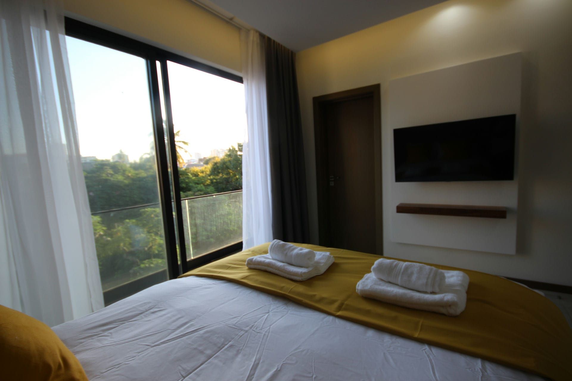 Bedroom with a large window, bed with yellow blanket and white towels, TV on wall.