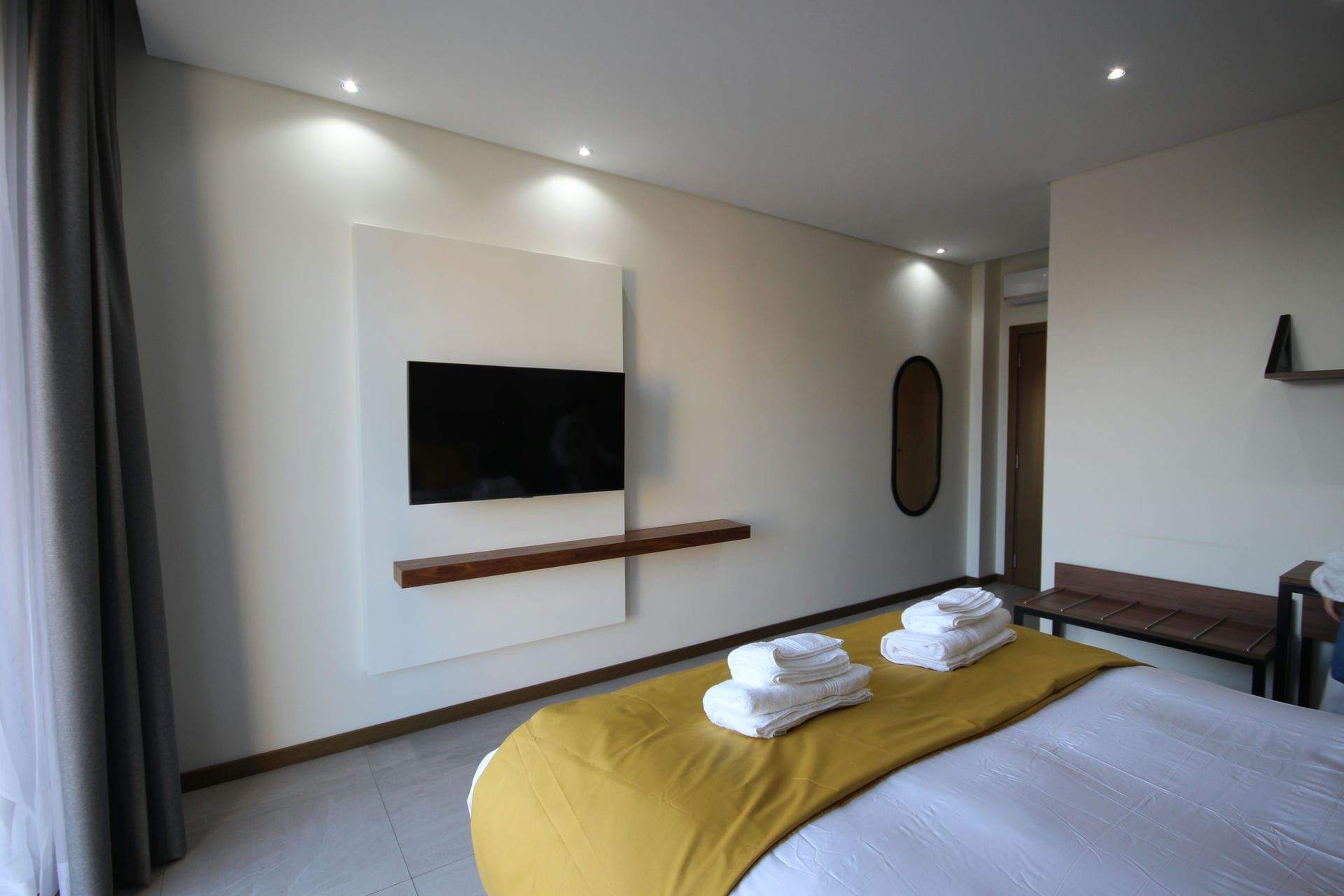 Bedroom with a large TV mounted on a white wall, bed with yellow blanket, white towels, and a wooden bench.