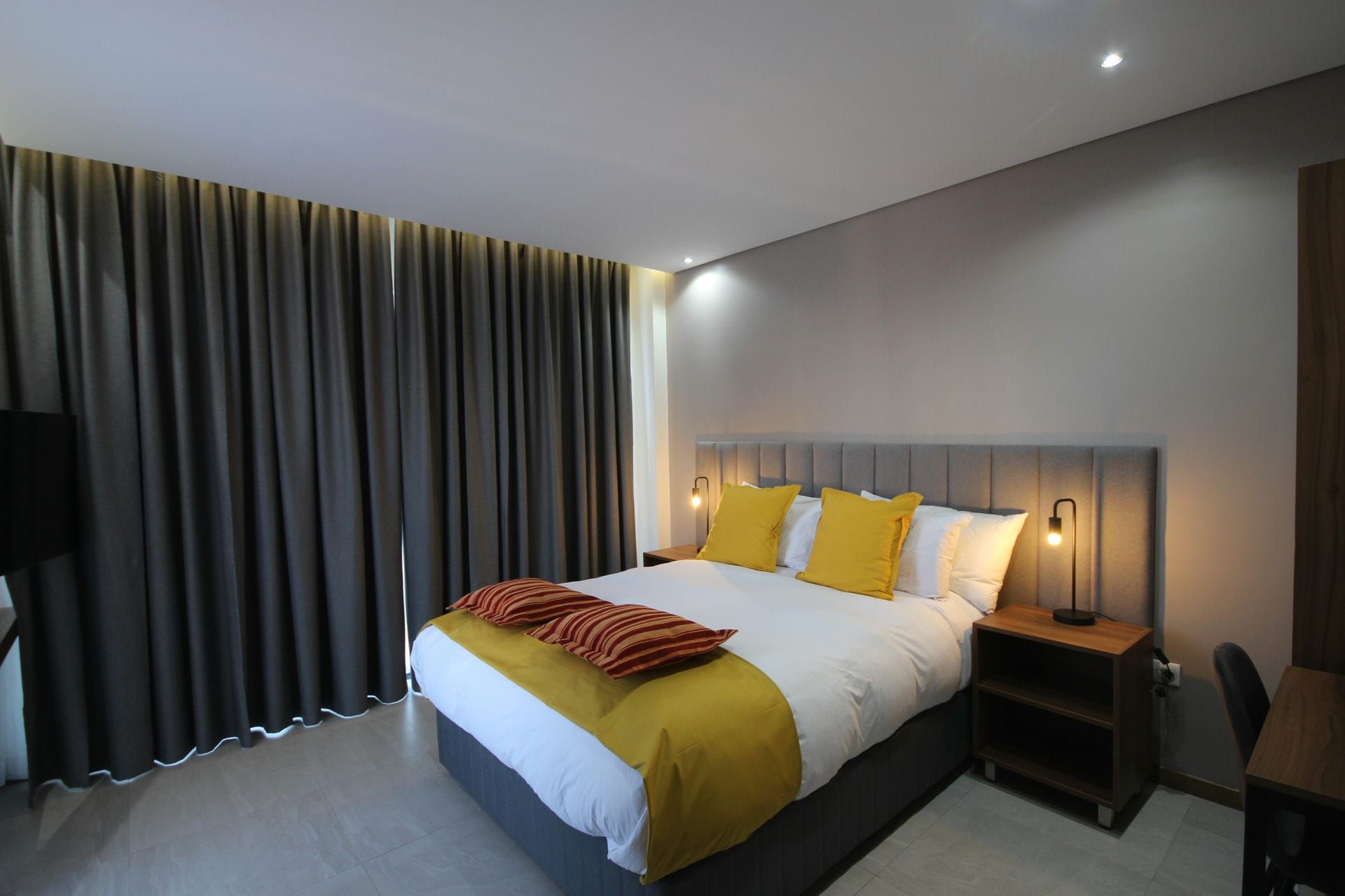 Bedroom with gray curtains, bed with yellow and orange pillows, nightstand with lamp.