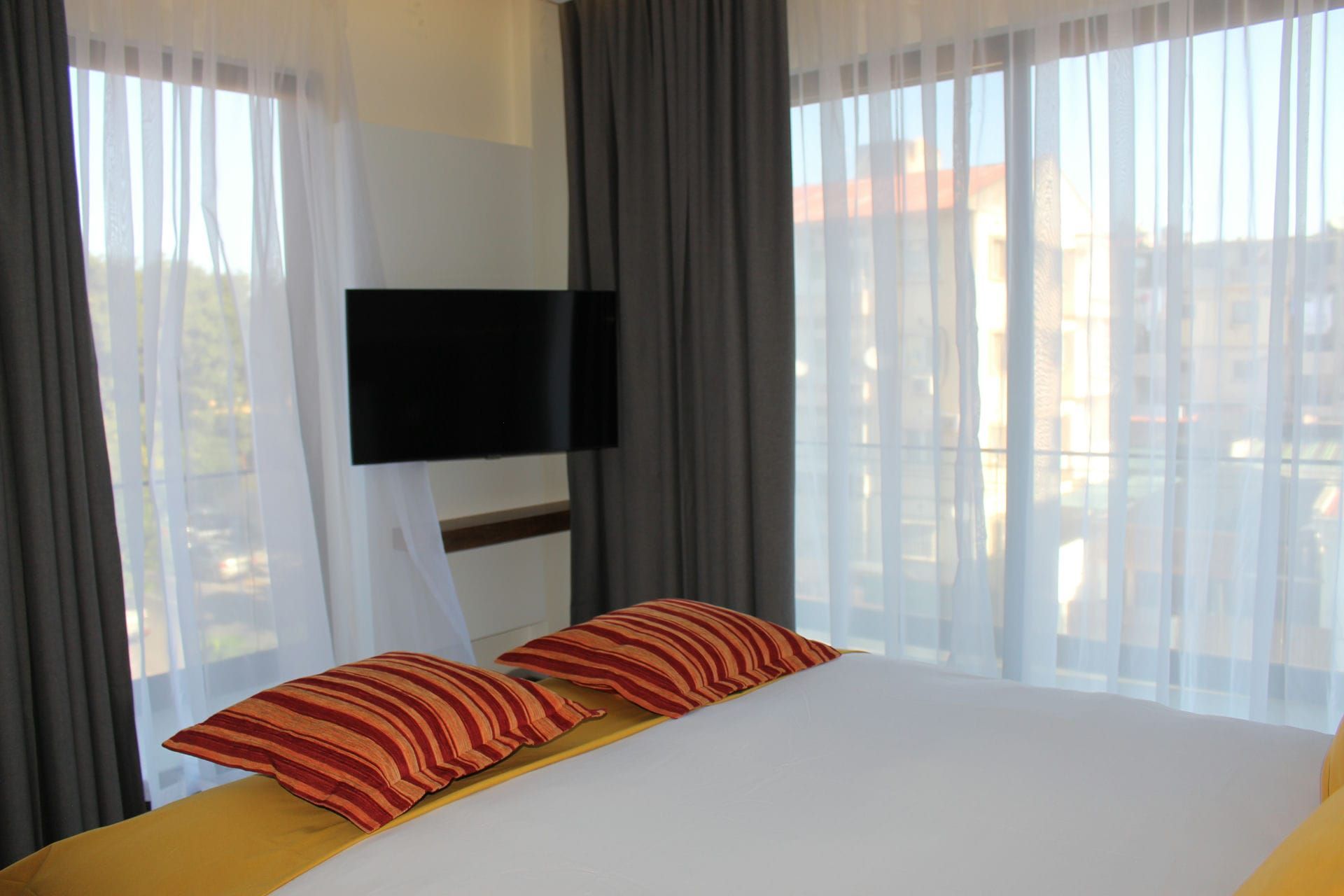 Bedroom with bed, pillows, TV on wall, sheer and gray curtains, and view.