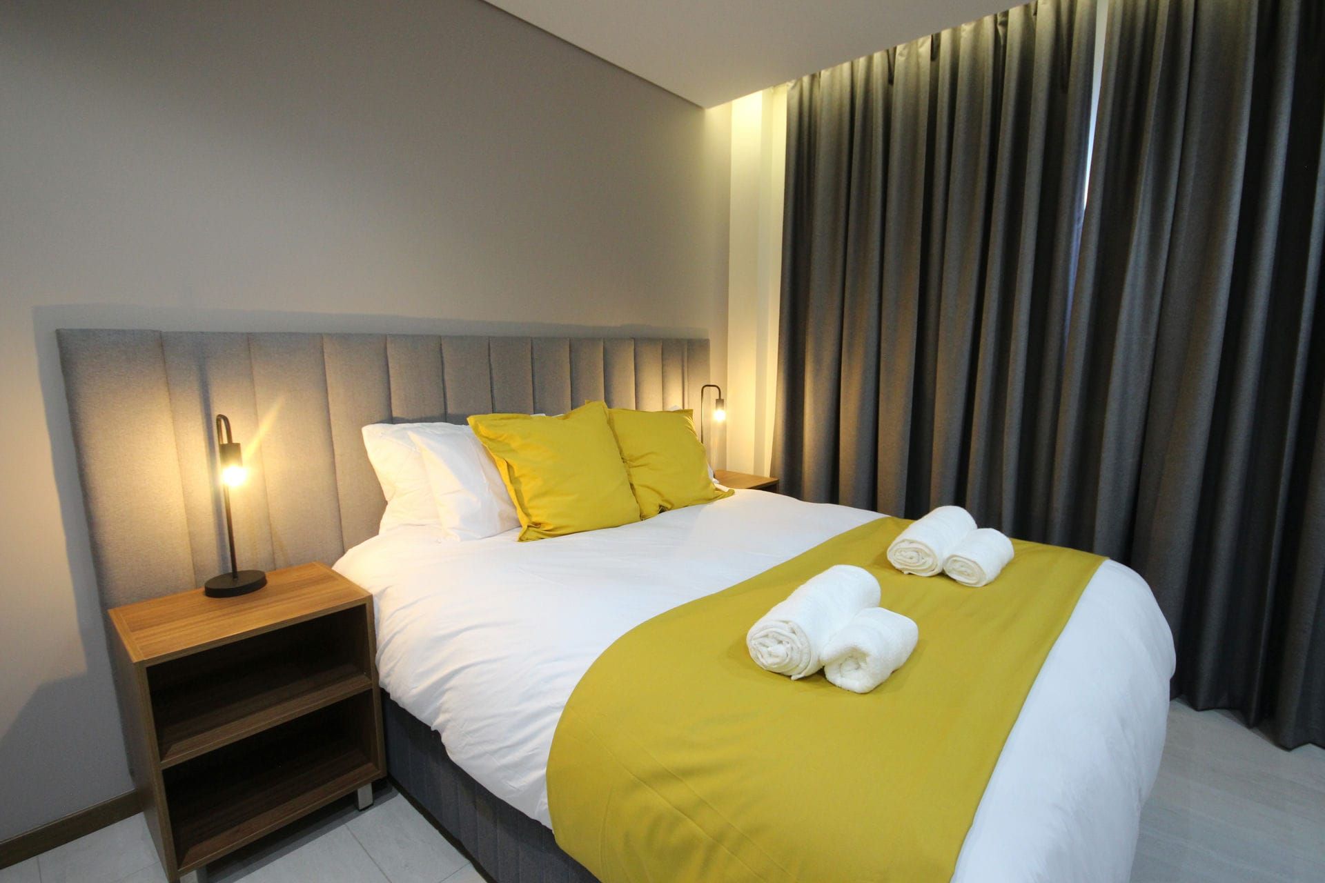 Bedroom with gray headboard, yellow and white bedding, wooden nightstand, and gray curtains.