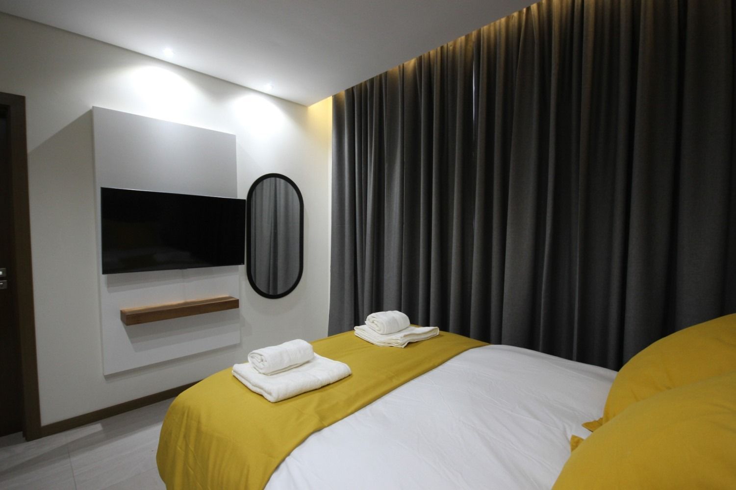 Bedroom with a bed, TV, and mirror. White and yellow bedspread, gray curtains, and a wooden shelf.