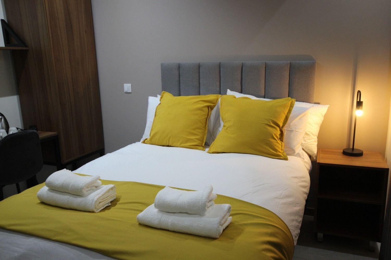 Bedroom with a gray headboard, yellow pillows, white bedding, towels, and a nightstand with lamp.