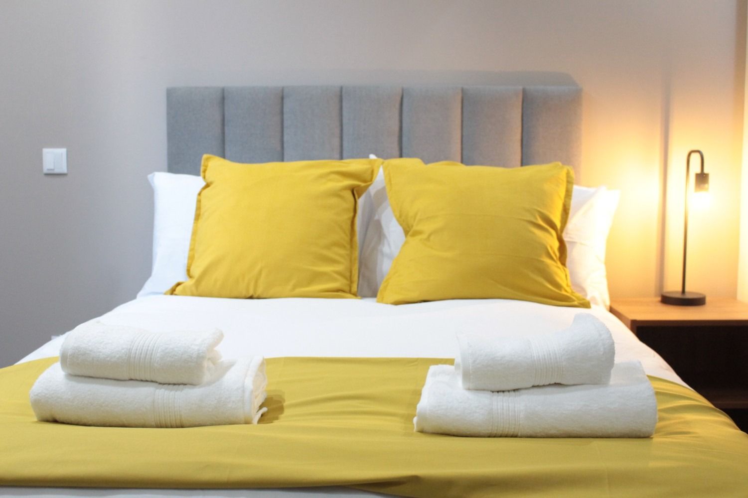 Bed with gray headboard, white linens, yellow pillows and throw, two folded white towels. Lit lamp on nightstand.