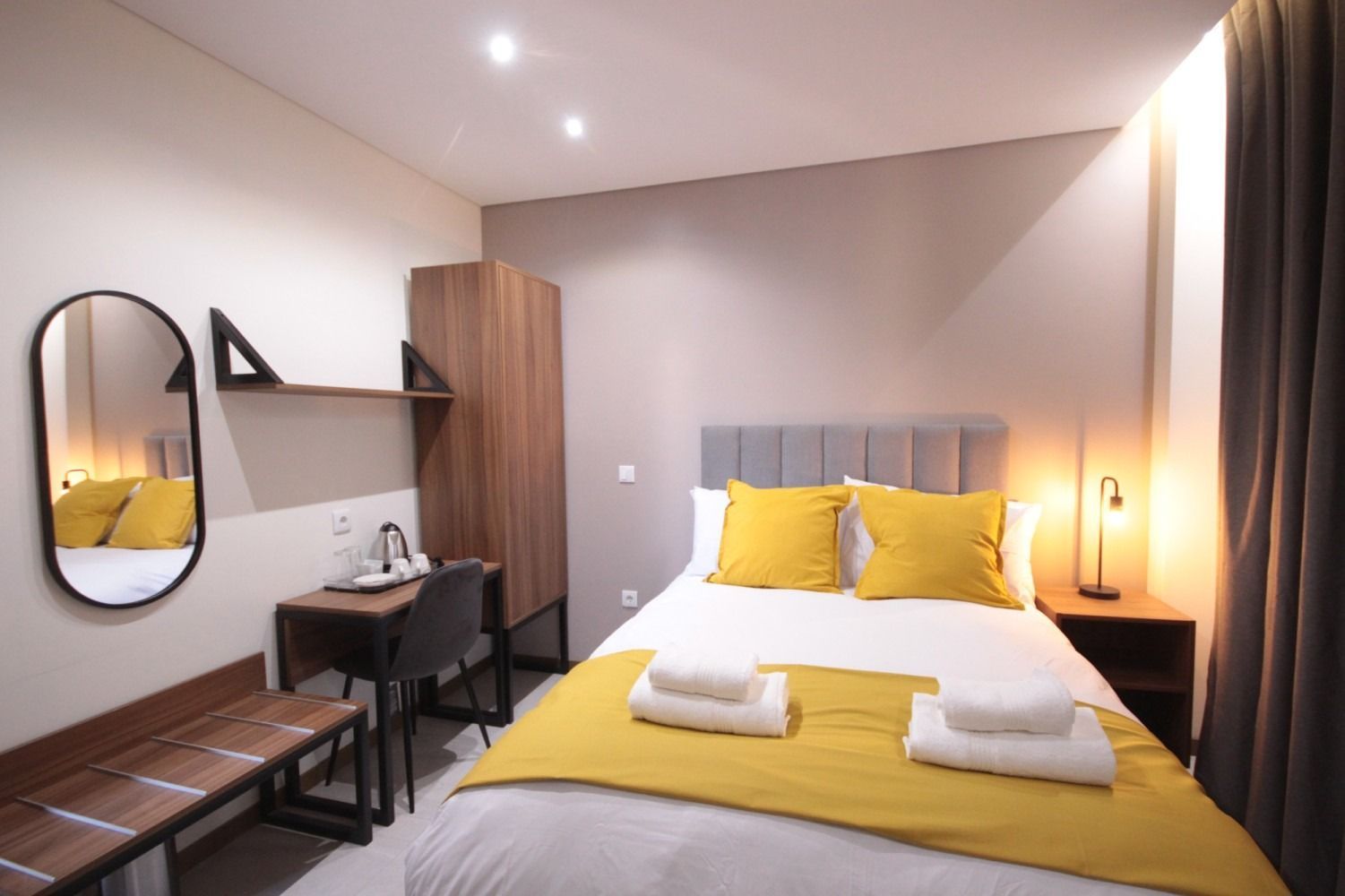 Hotel bedroom with a bed, desk, mirror, and wardrobe, featuring yellow and gray accents.