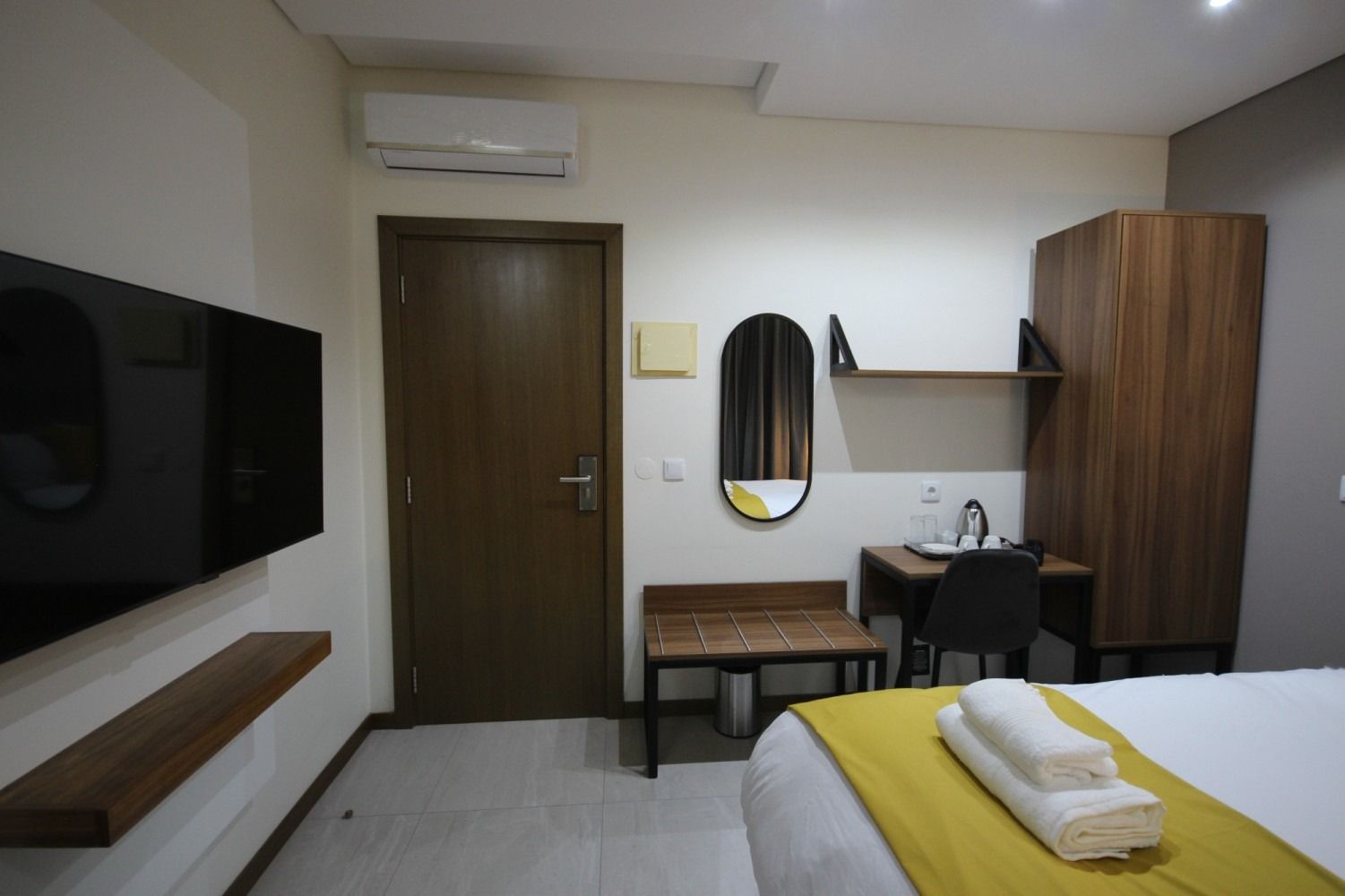 Hotel room interior with a bed, desk, wardrobe, and mounted TV.