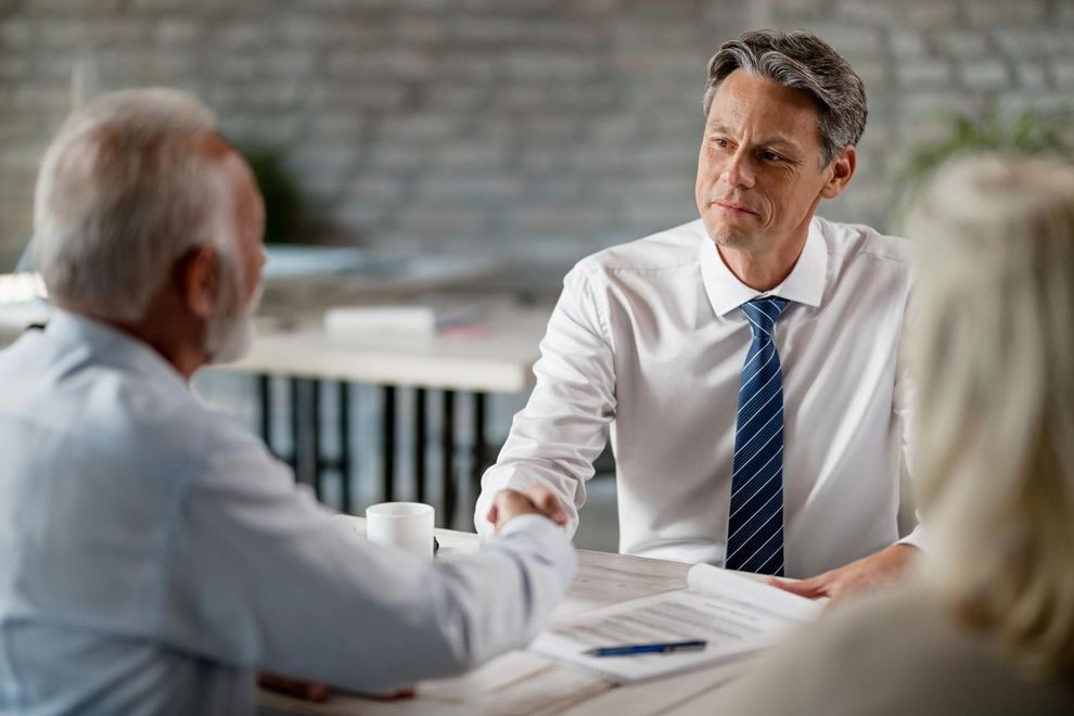 Man in white shirt shakes hands with older man at a table, woman seated nearby.