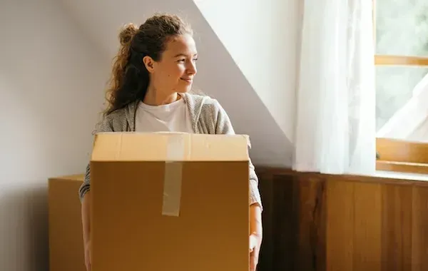 Woman carrying a cardboard box, looking out a window in a room with wood trim.