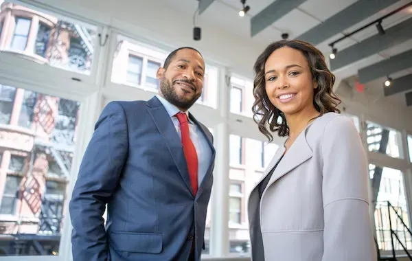 Two professionals smiling, one in a suit with a red tie, the other in a light gray blazer, standing near large windows.
