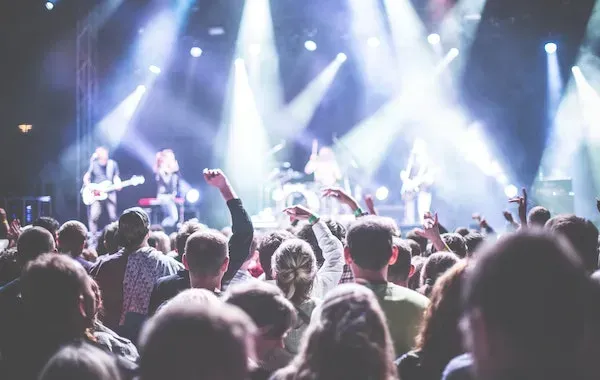 Concert crowd with raised arms, facing a band on stage under bright stage lights.