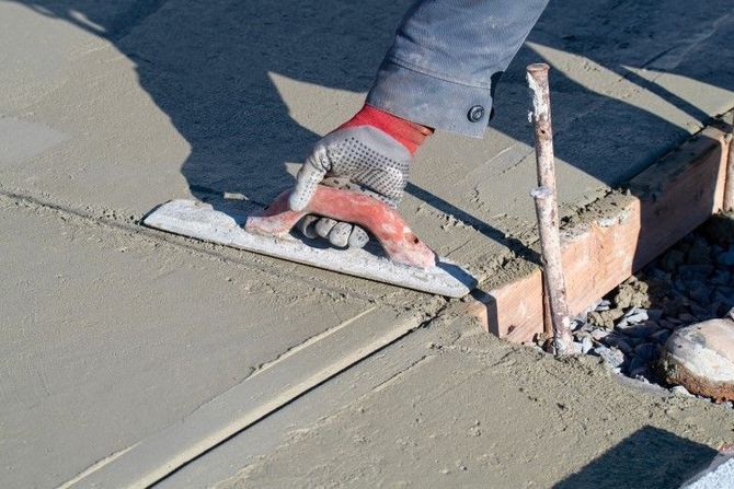 Worker smoothing wet concrete beside a wooden form and sidewalk edge.