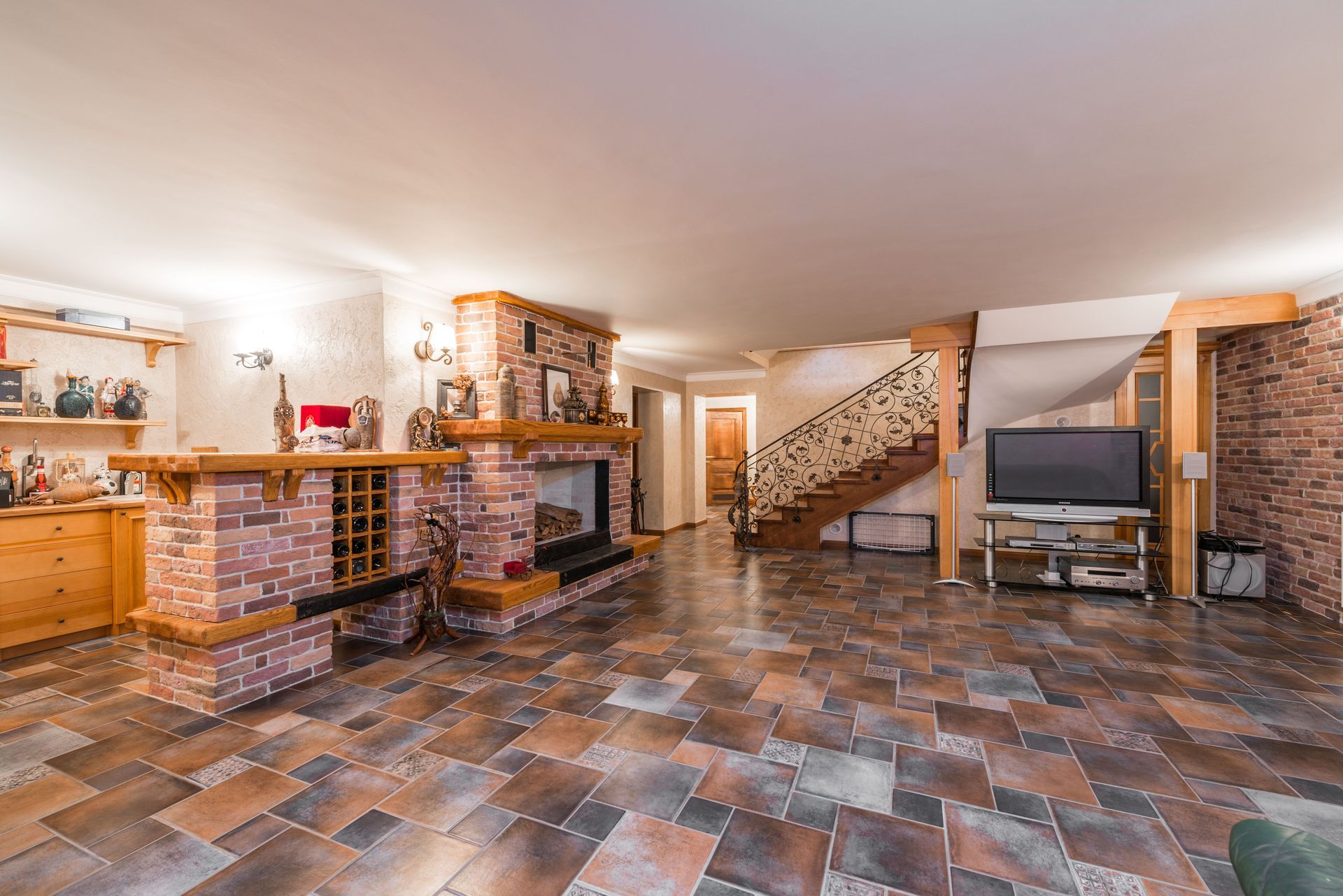 Basement rec room with brick bar, tiled floor, TV, and staircase under warm lighting
