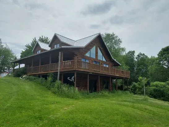 Wooden cabin with wraparound porch on a grassy hill under a cloudy sky.
