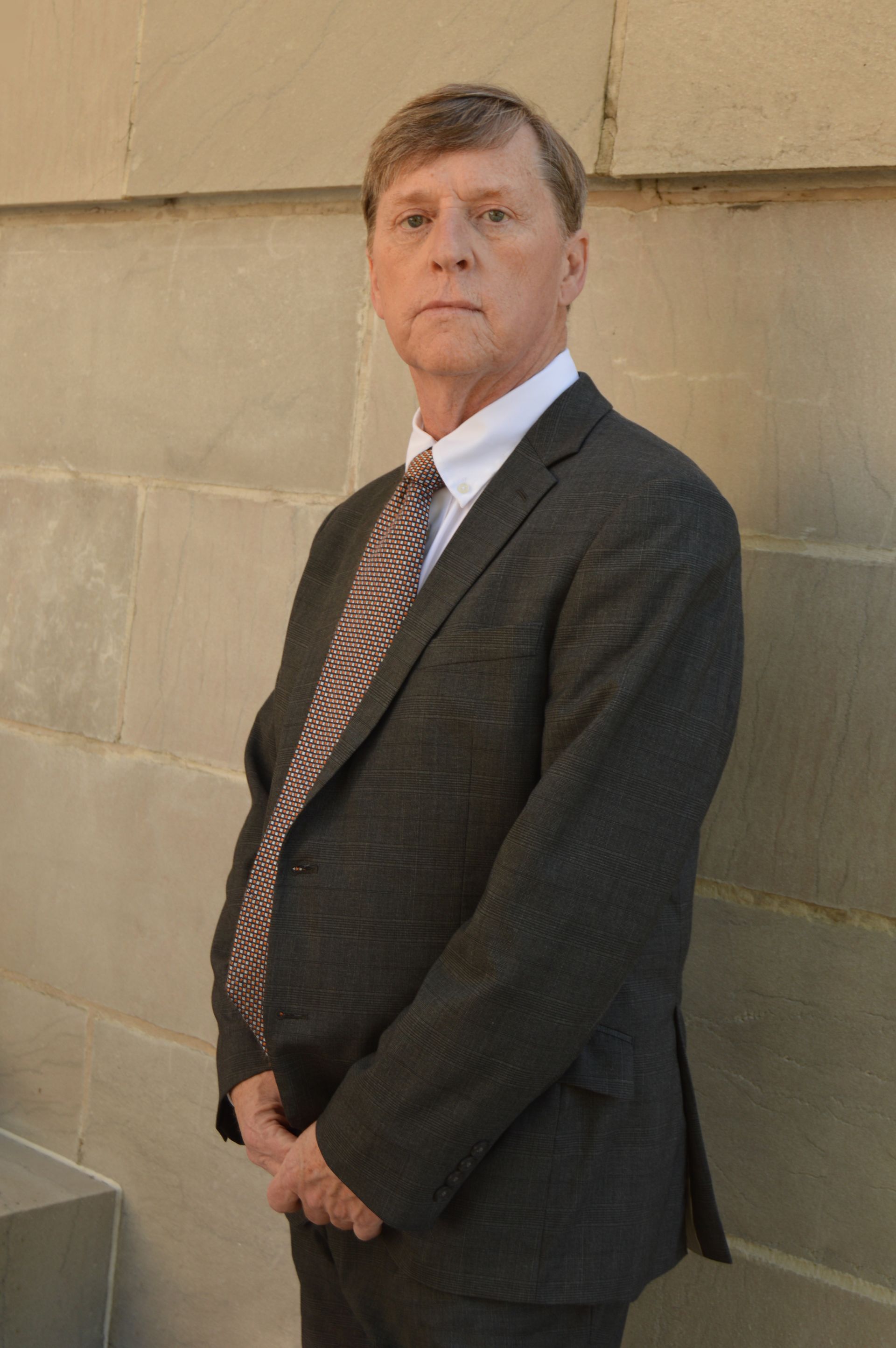 Man in a suit and tie standing against a light-colored stone wall, arms crossed.