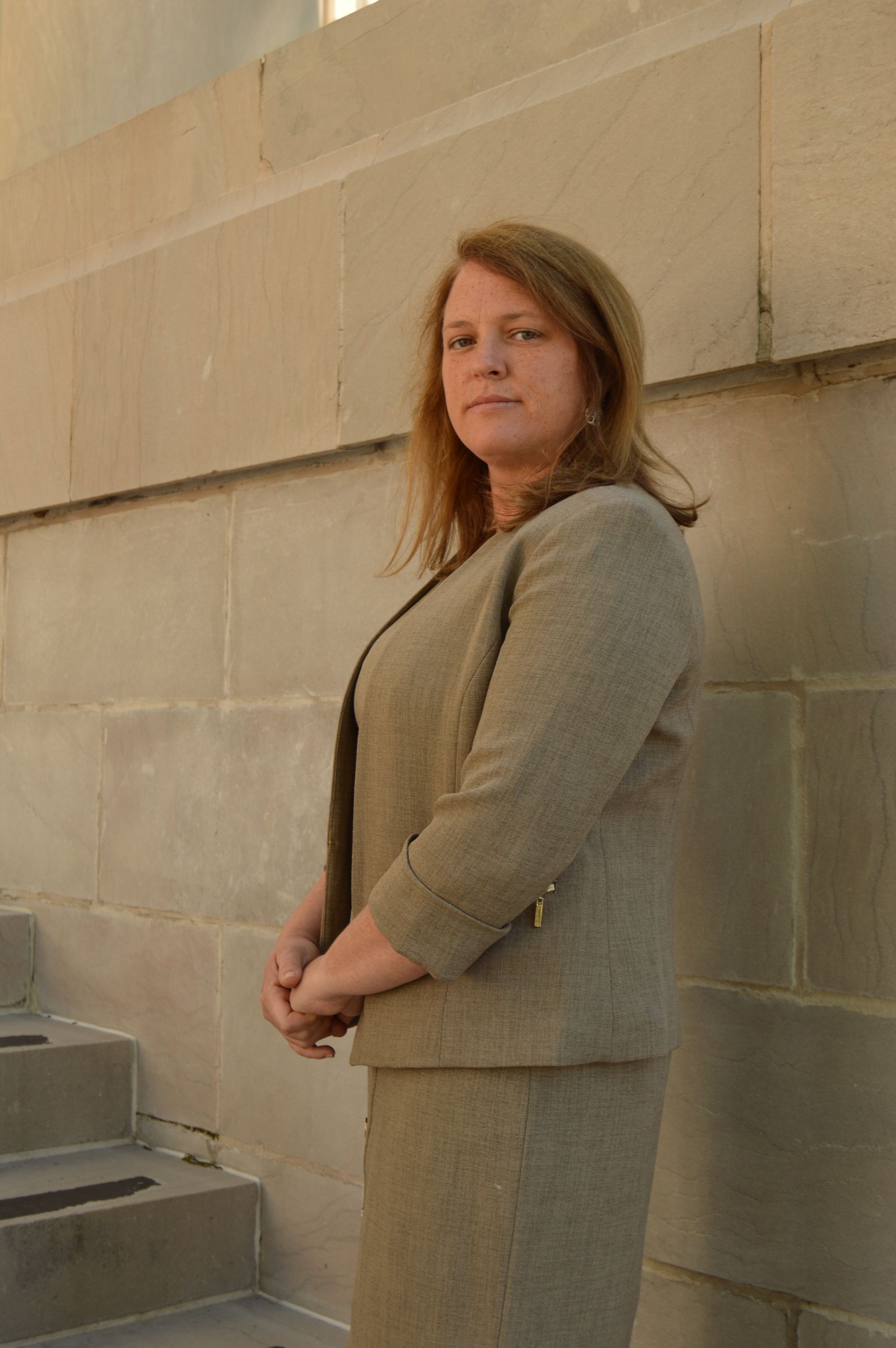 Woman in a tan suit standing by a stone wall, arms crossed, looking at the camera.
