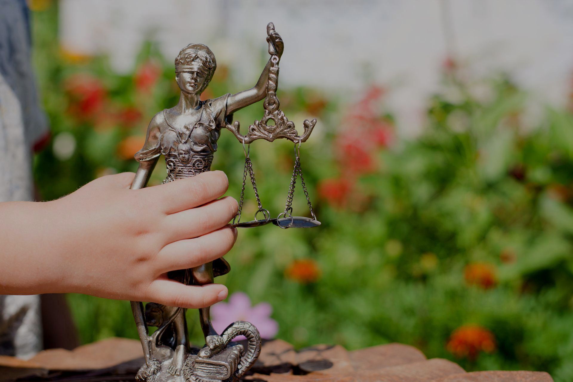 Child's hand holding a statue of Lady Justice with scales, outdoors with blurred flowers in background.