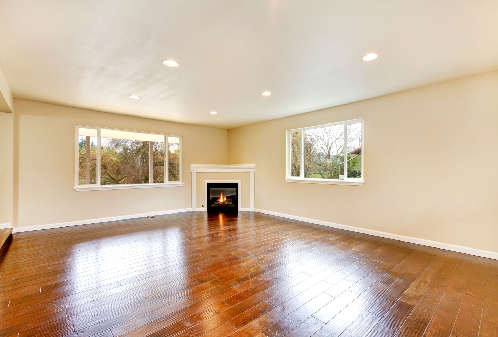 A bright, empty living room with light beige walls, dark hardwood floors, and a corner fireplace with a lit fire.