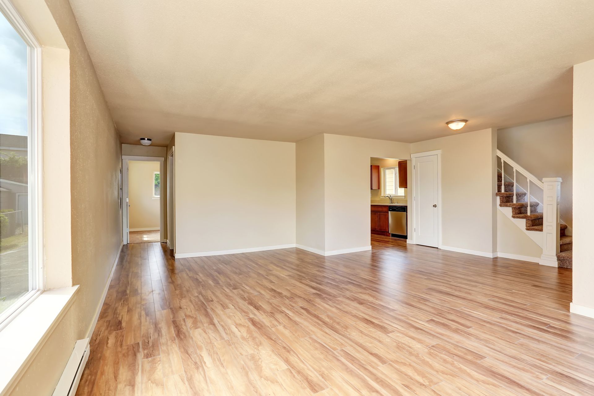 An empty, well-lit living area featuring hardwood floors, beige walls, a side staircase, and a view into a kitchen.